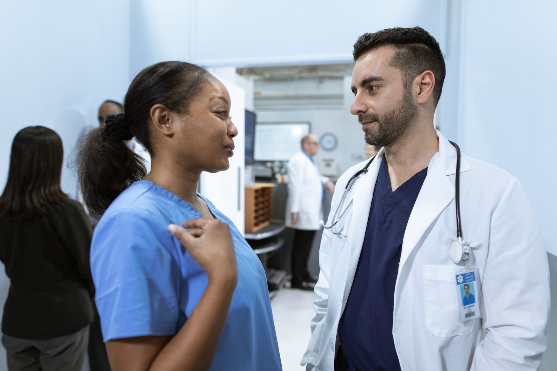 Black female nurse in blue scrubs talking to male physician wearing white lab coat.