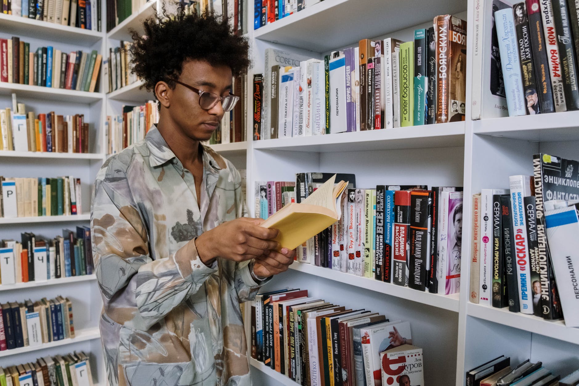 Black man with glasses and curly hair reading book in library.