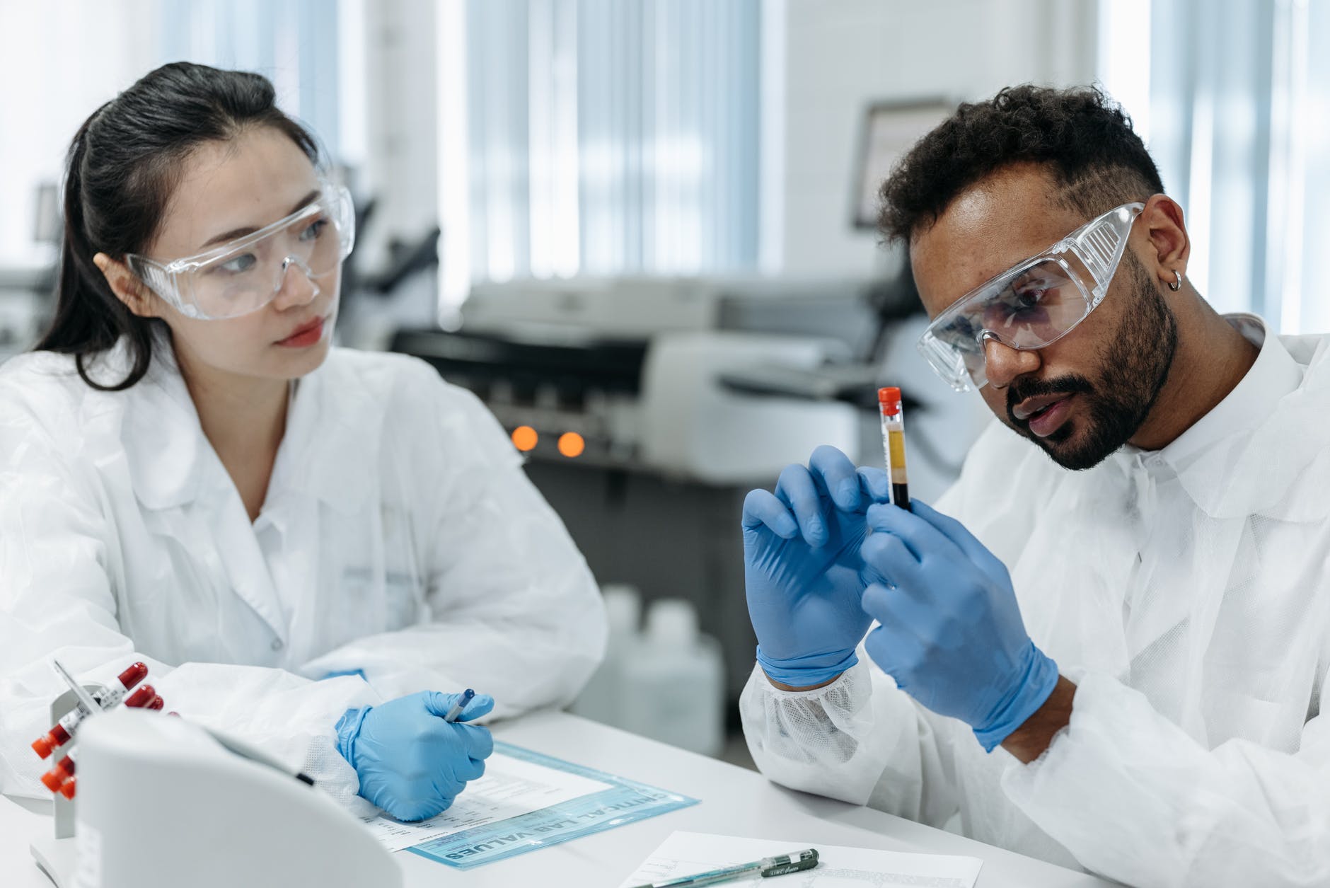 Black man holding tub with red stopper and wearing lab coat, blue gloves, and eye protection. Asian woman taking notes also wearing disposable lab coat.
