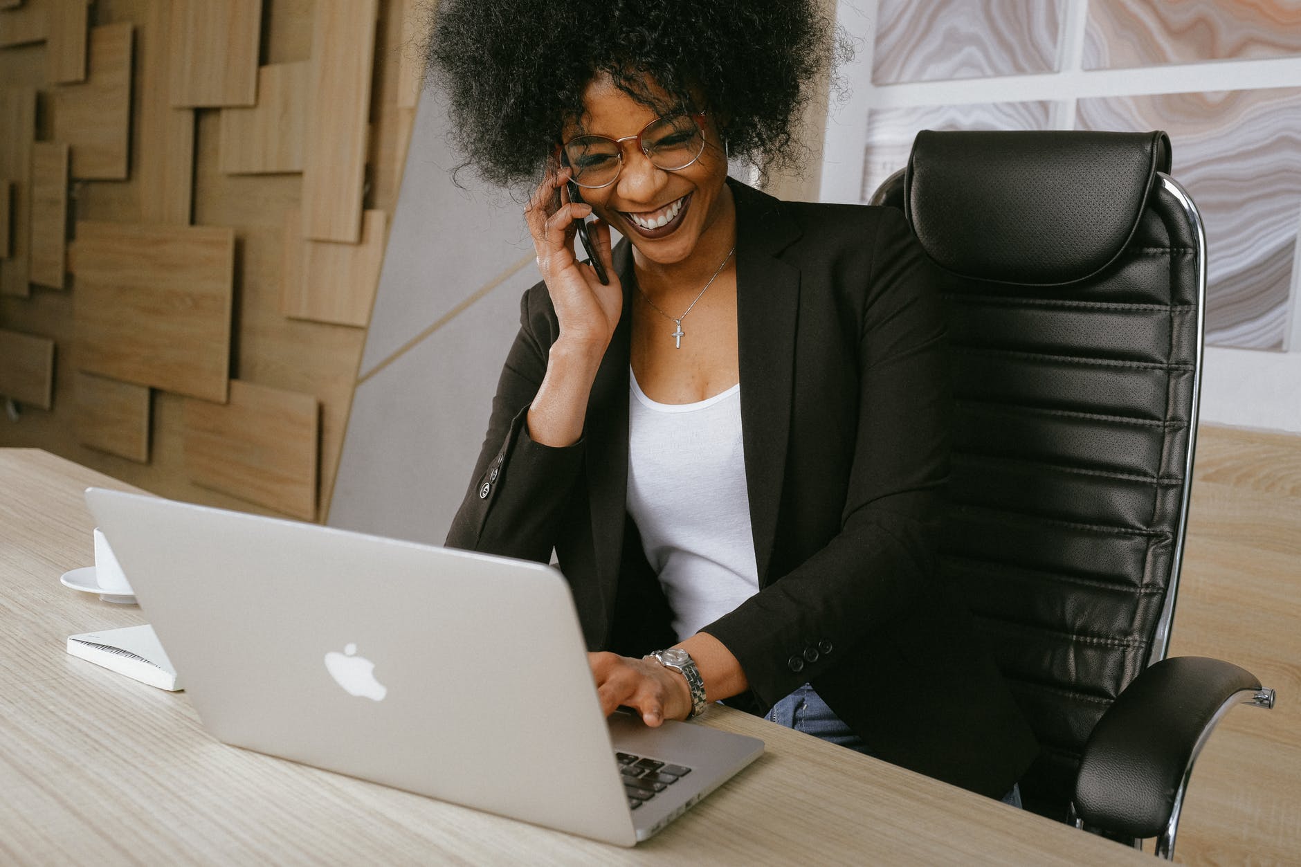 Black woman on cell phone and working on laptop at desk.