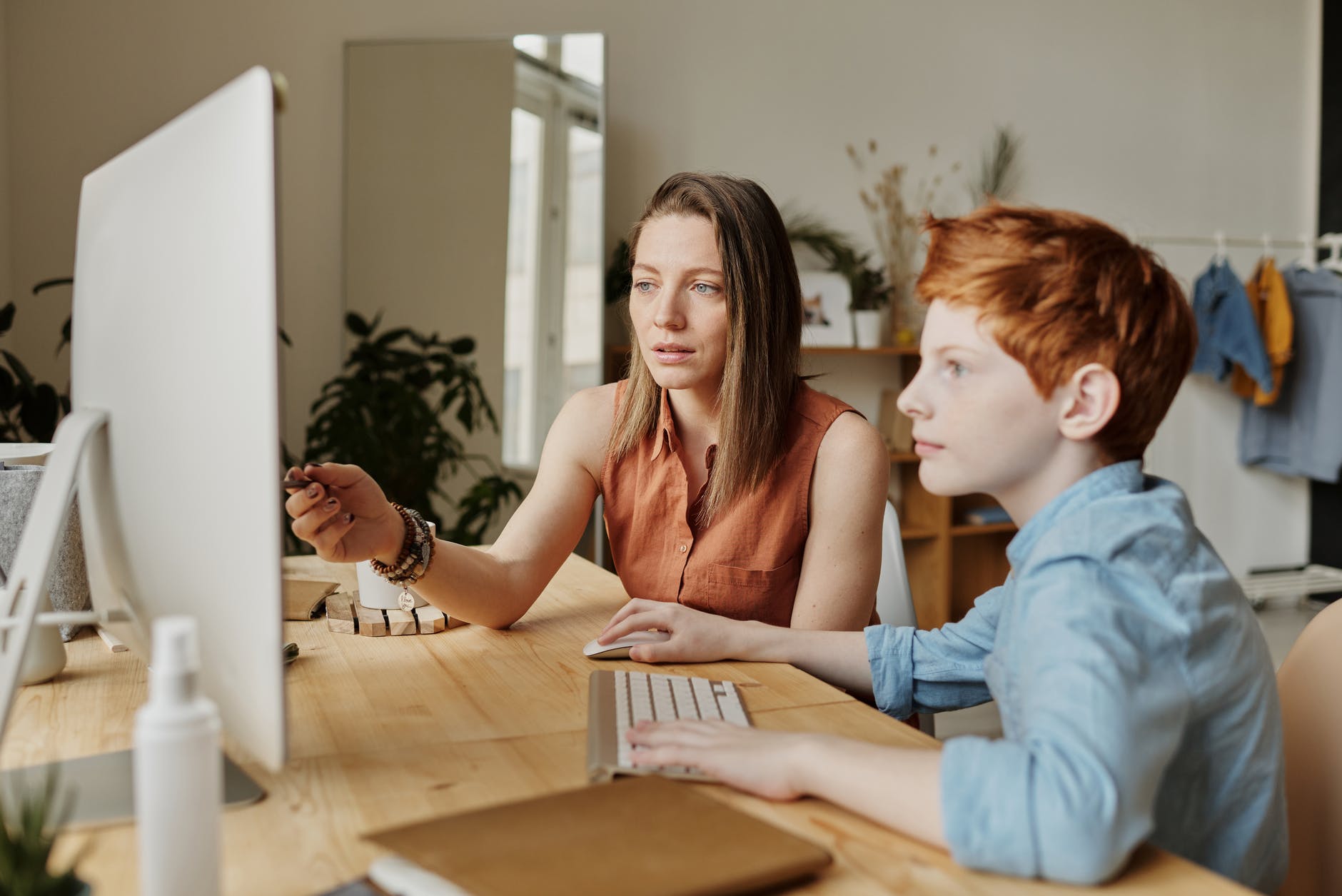 White teacher with brown shoulder-length hair pointing at computer screen. Red haired boy is at the keyboard.