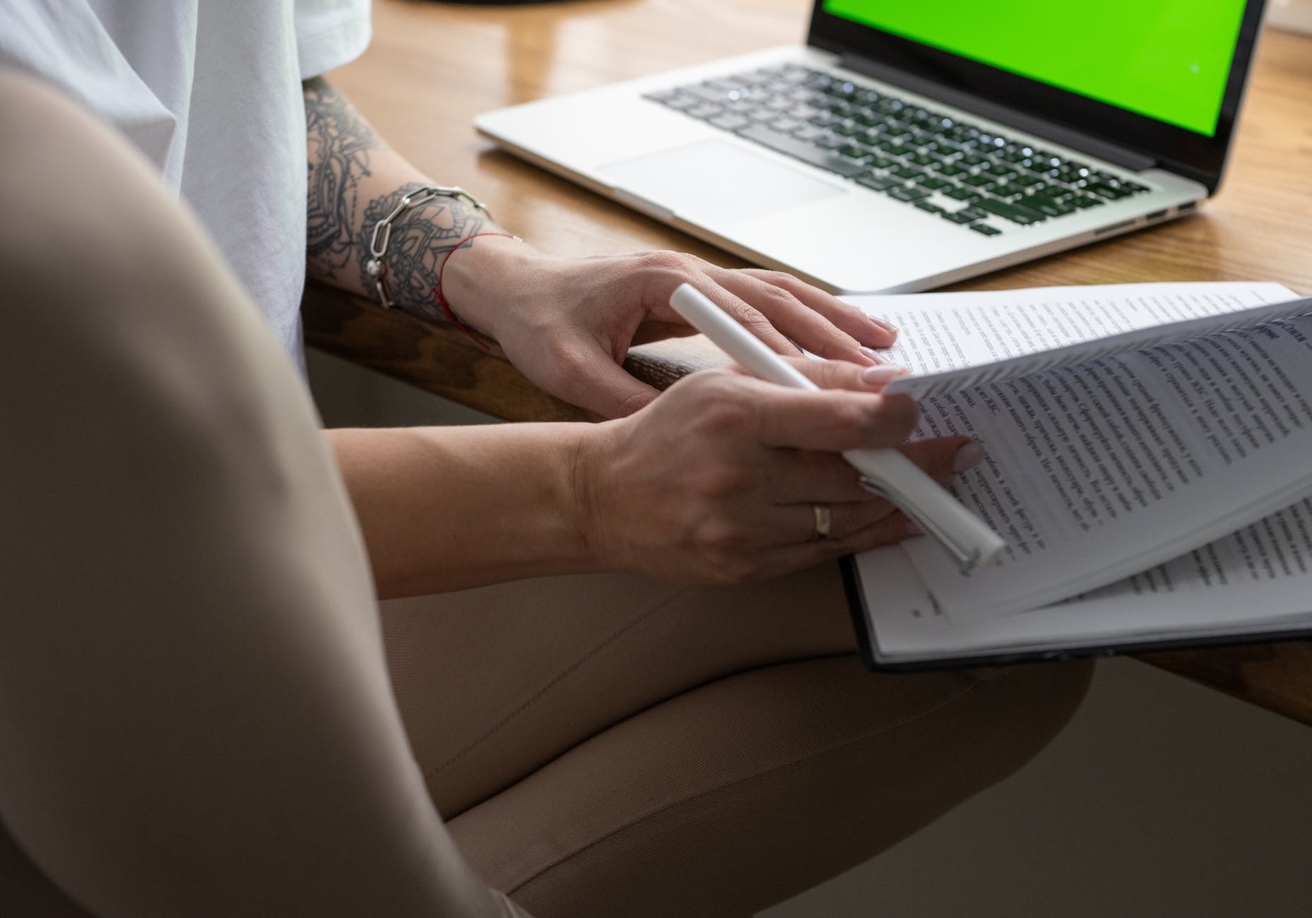 Hands with tattoos holding textbook and working on laptop with green screen.