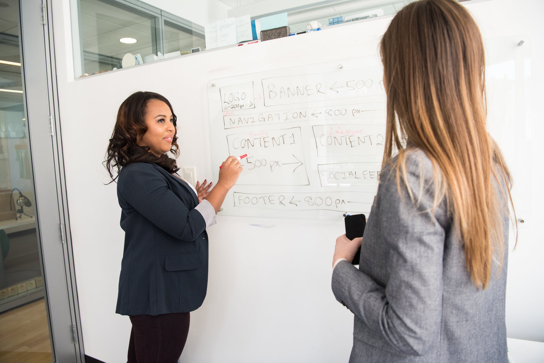 Black woman teaching at whiteboard to another person facing the whiteboard.