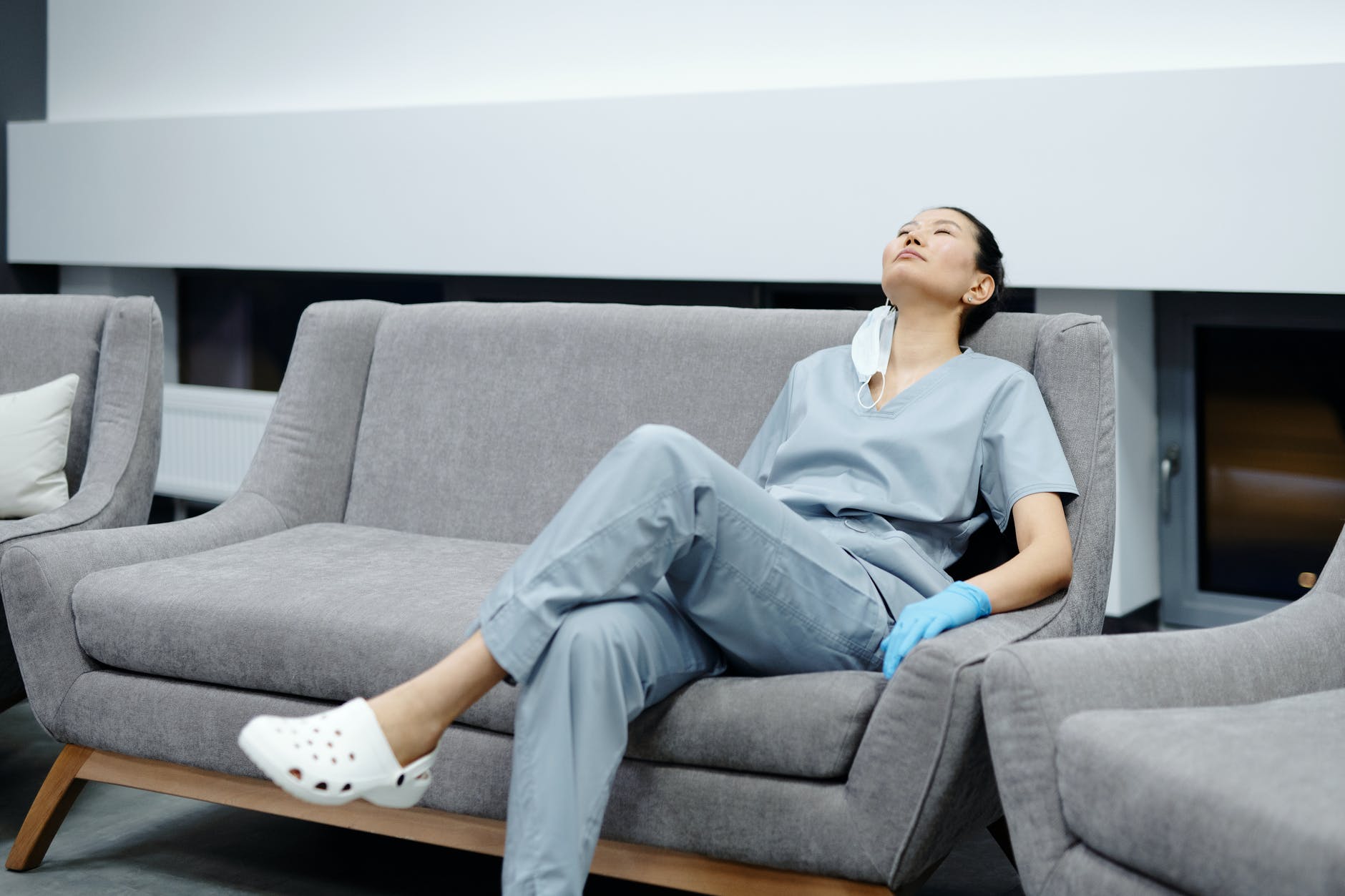Female asian nurse sitting back on couch wearing scrubs and gloves.