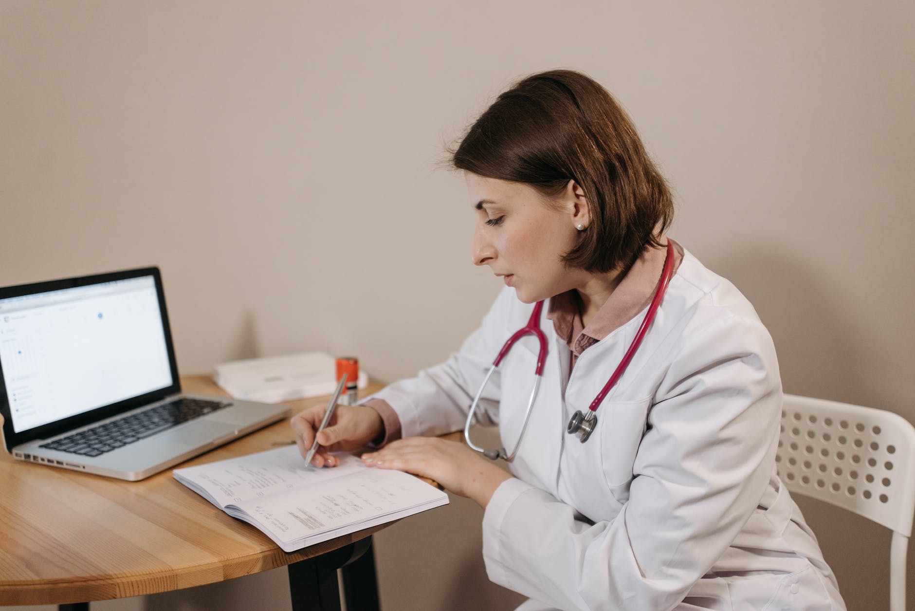 Female clinician writing a prescription in a book next to open laptop.