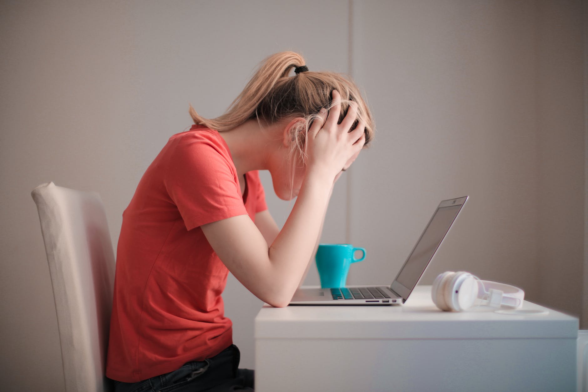 Woman with blond ponytail and hands on forehead holding head seated at desk with laptop and blue mug.
