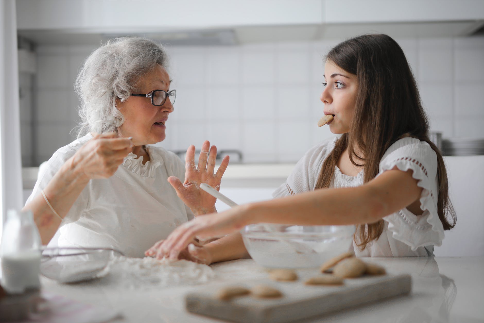 Older woman with gray hair sharing cookies with younger woman. Woman has long brown hair and cookie in mouth.
