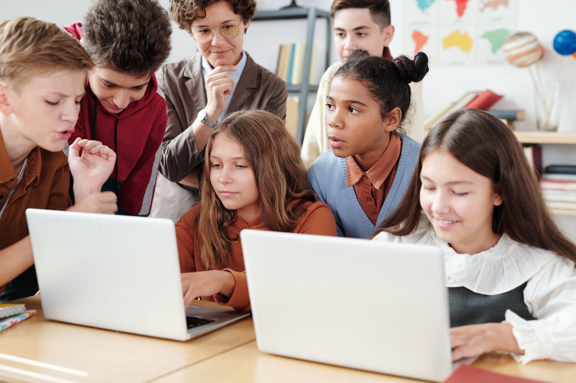 Group of kids in front of two laptops. Teacher is woman with glasses behind kids.