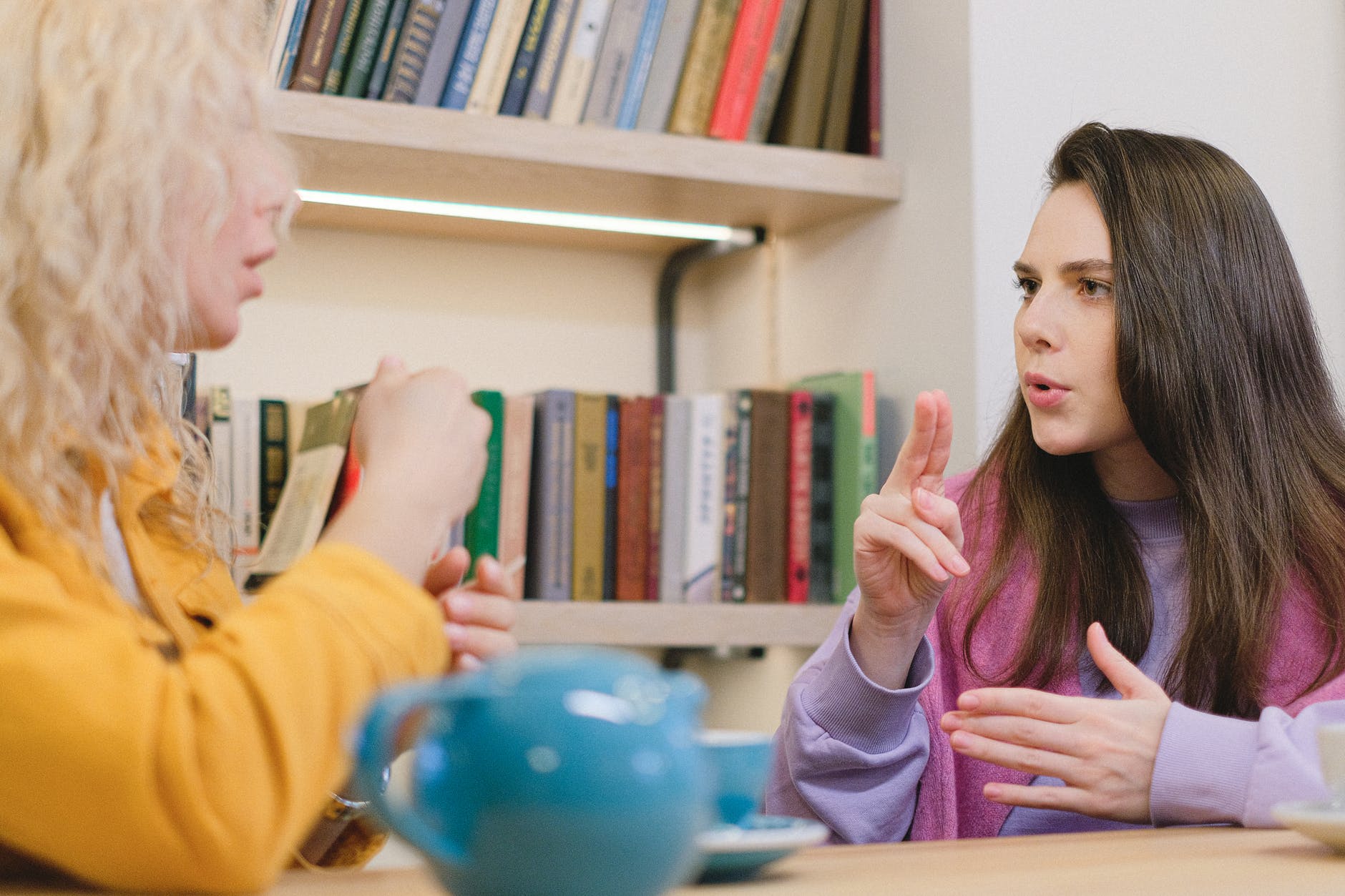 A woman facing the camera and communicating with sign language. They have long brown hair. They are facing a second woman with curly blond hair.