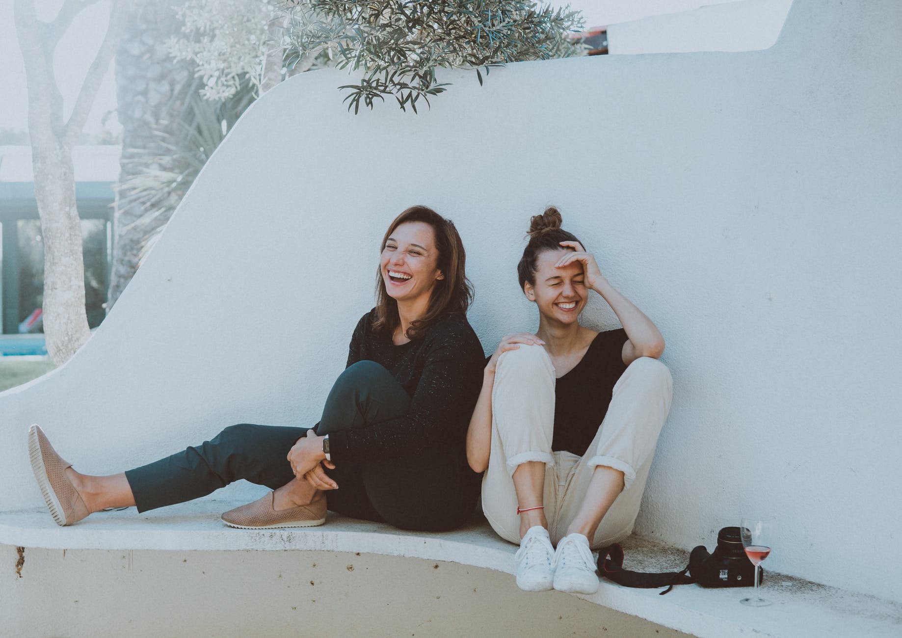 two women sitting down against white wall laughing