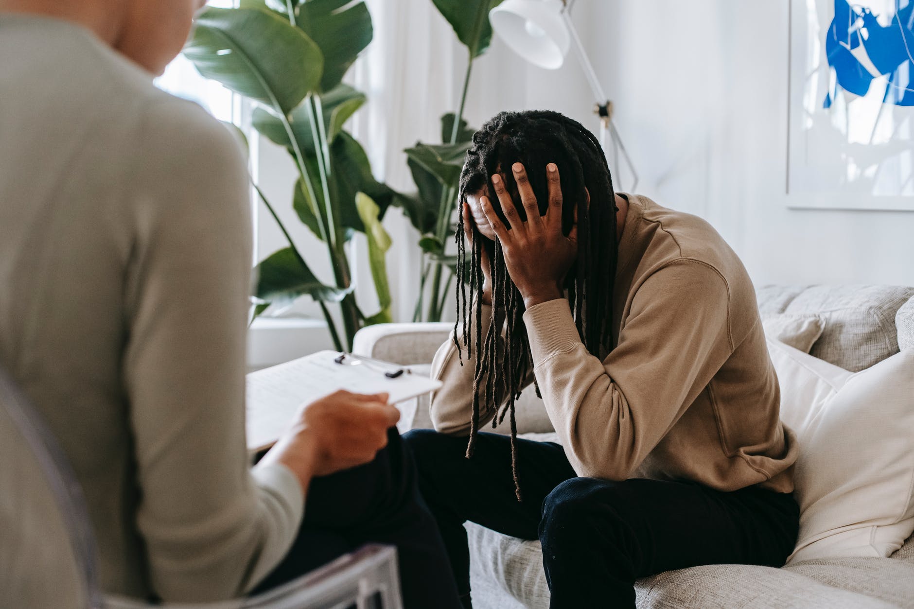 Black man with long hair in dreads on couch with hands on forehead holding head. Person is facing them with clipboard