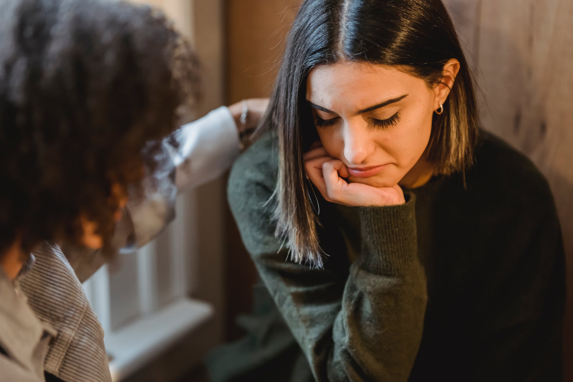 woman with brown shoulder-length hair talking to person with curly hair. Woman seems concerned, with hand holding head.
