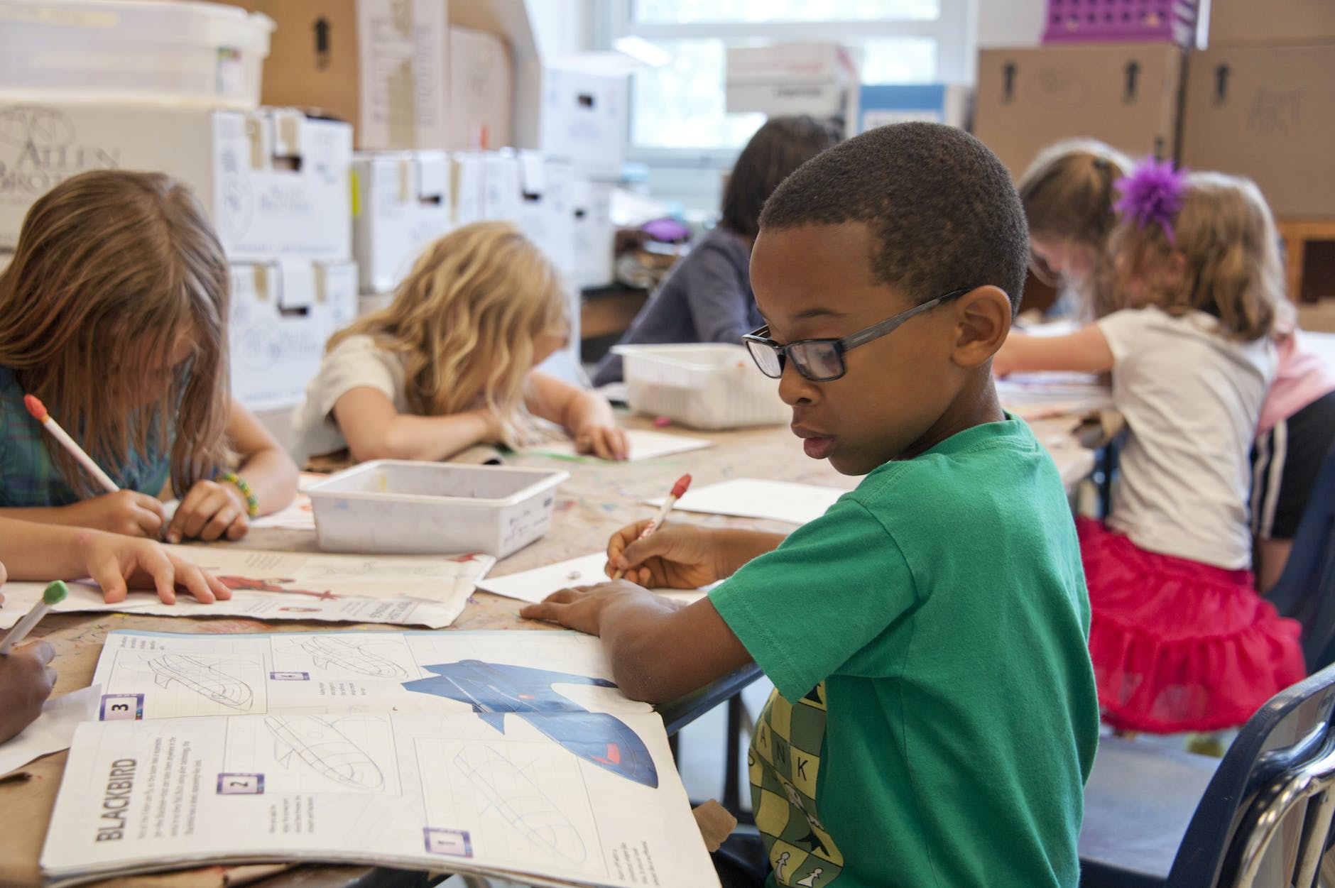 Black boy surrounded by other children working around a long table on writing on paper.