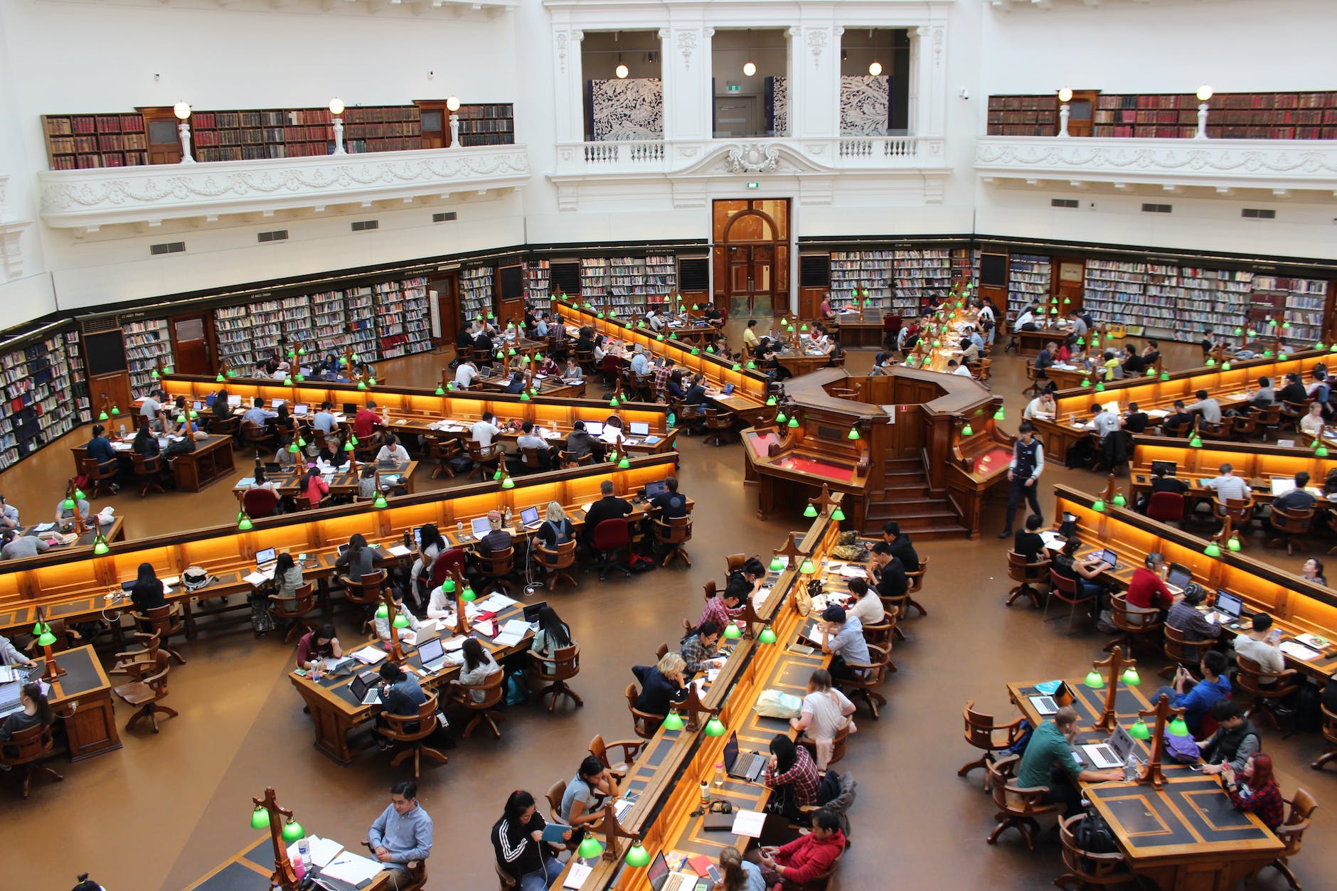 rows of long tables in star pattern in library. Numerous people are seated reading and there is a center desk in the middle. Bookcases cover the walls of the room.