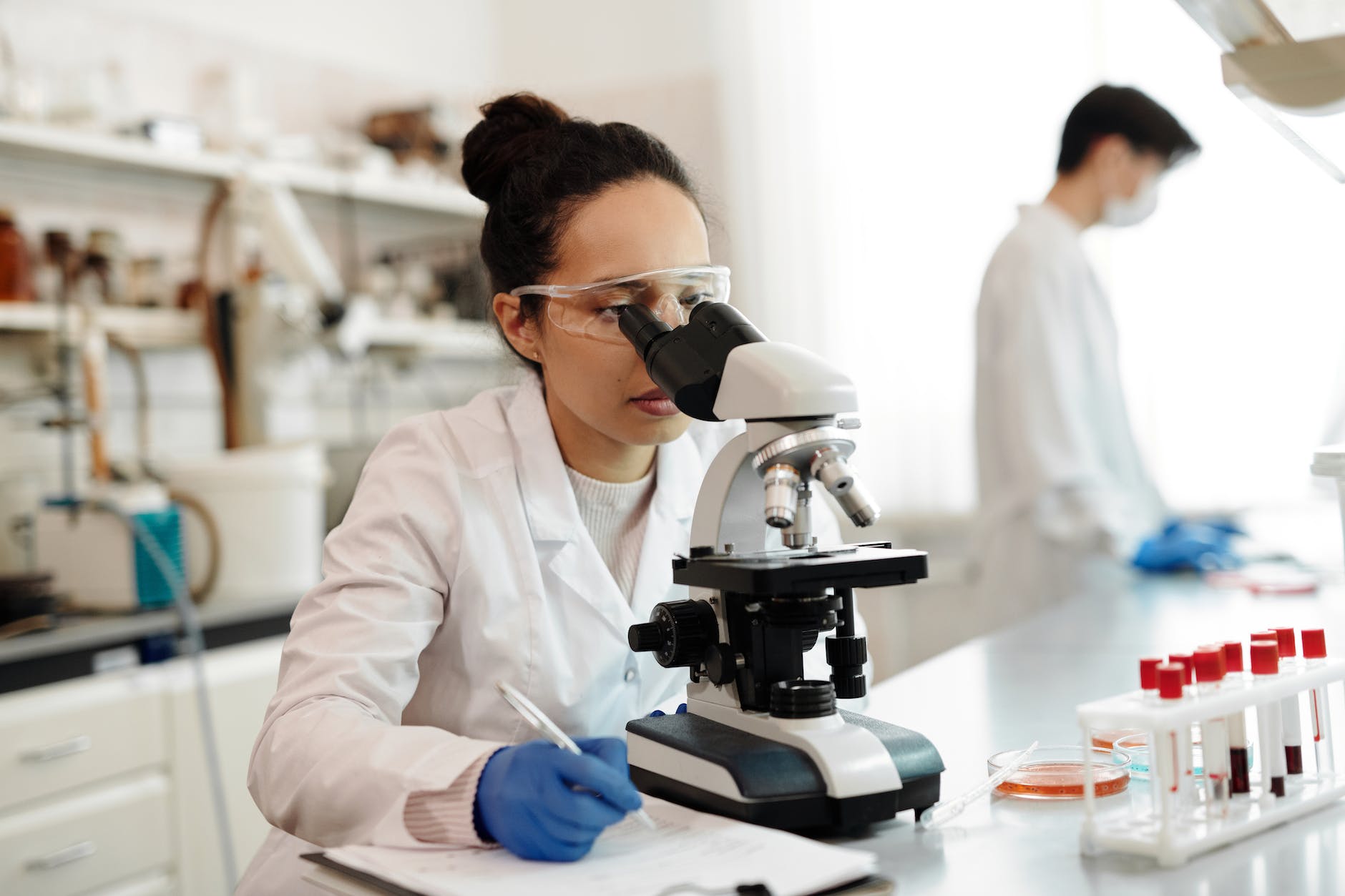 Woman wearing a white lab coat and blue gloves and safety glasses working a microscope. The blurred person behind standing up and wearing white lab coat.