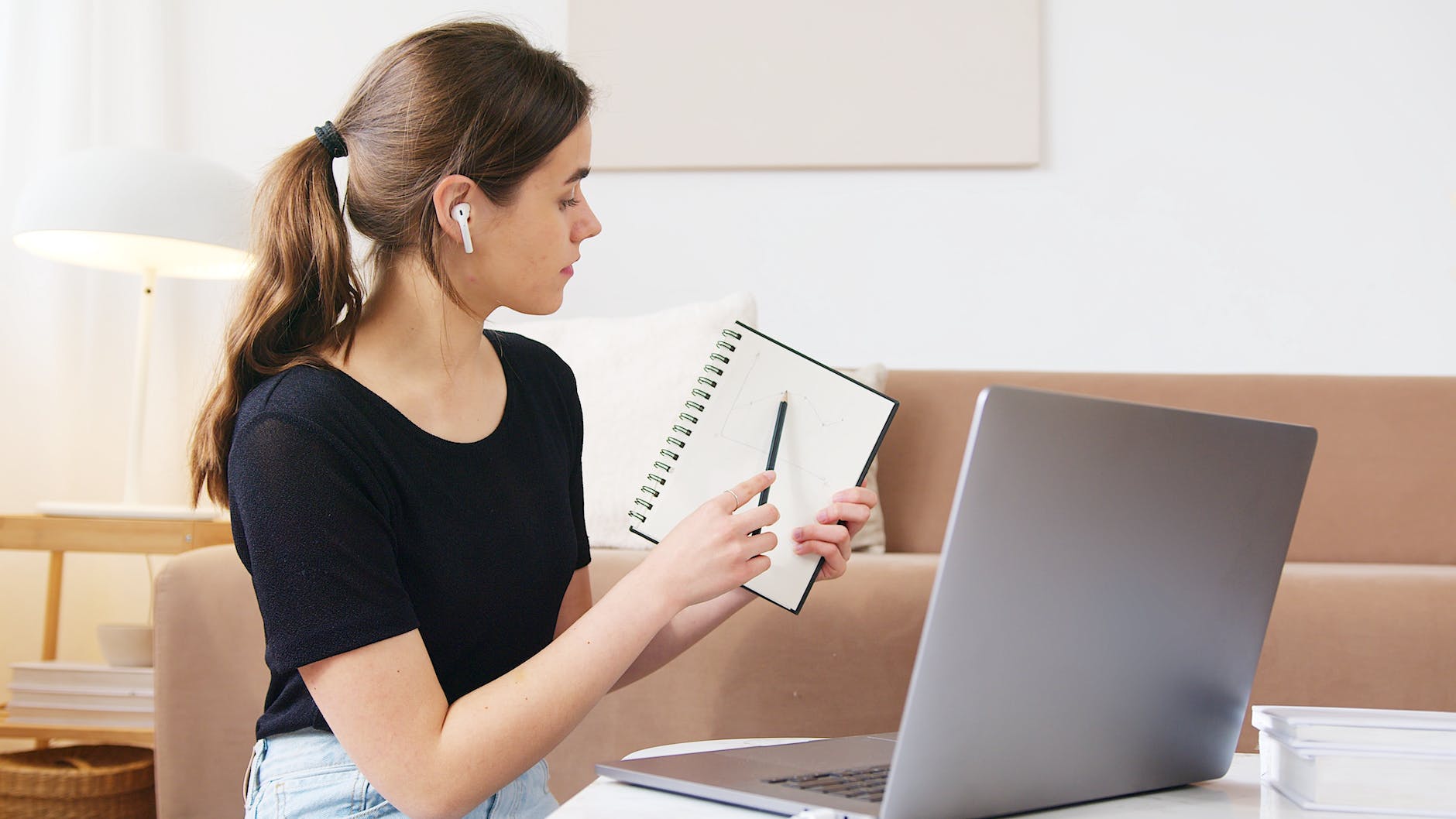 White woman with brown hair wearing earbuds holding pencil and pointing to notepad in front of open laptop