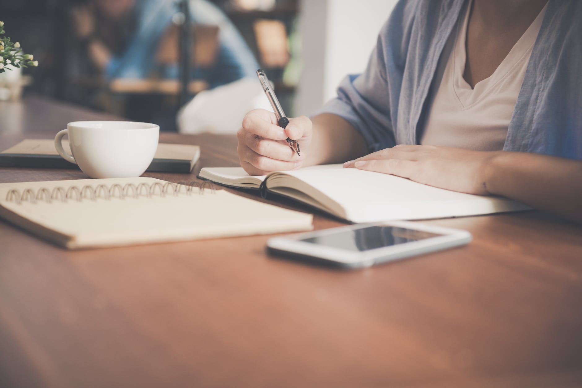 person writing in a notebook next to teacup and tablet
